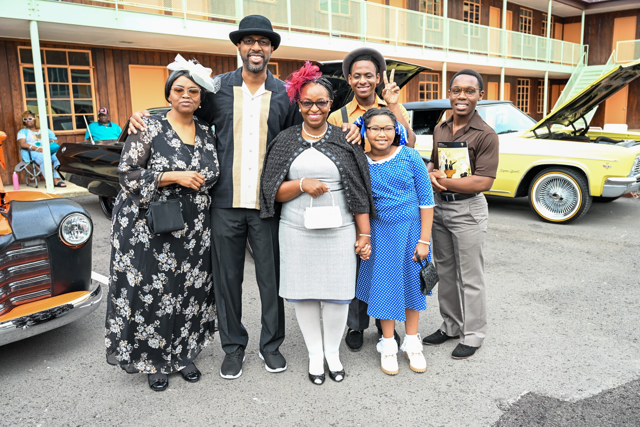 Six family members dressed in 1960's clothing stand in front of cars from the 1960's. The Gaston motel is seen in the background.