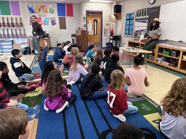 A ranger is talking in front of a group of about 20 seated students.