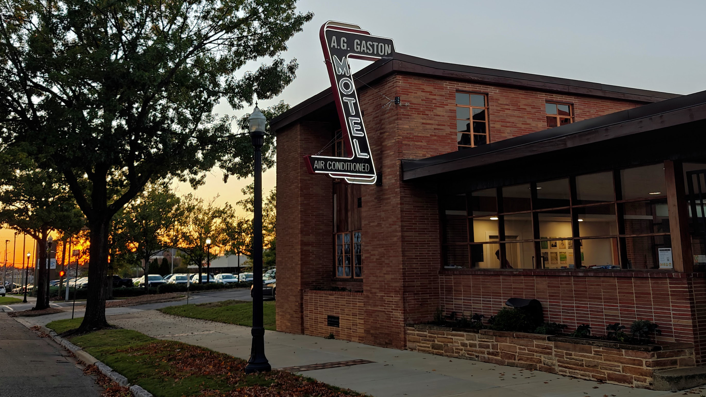 An illuminated neon sign reading A.G. Gaston Motel hangs from a two story brick building. A sunset is in the background.