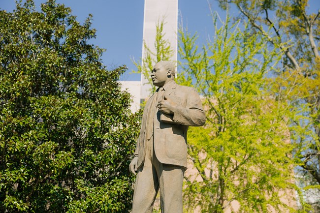 A statue of a man on a pedistal. Behind are trees and a building