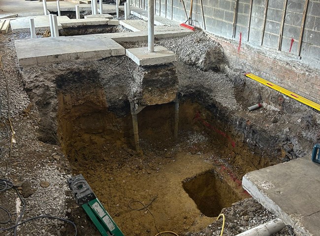 A rectangular clay pit about 6 foot deep sits with a smaller deeper square hole inside the larger hole. Metal supports rise from the pit to support the ceiling.