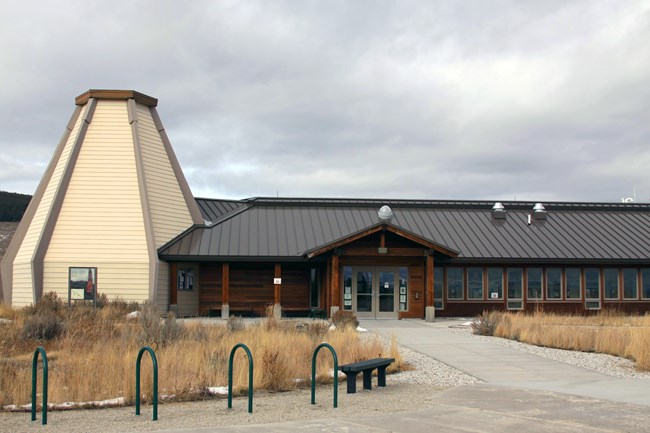 The Visitor Center at Big Hole National Battlefield, a building with a distinctive, tall, angular structure resembling a tipi next to the main entrance, with a metal roof and large windows, set against a cloudy sky and surrounded by natural grasses.