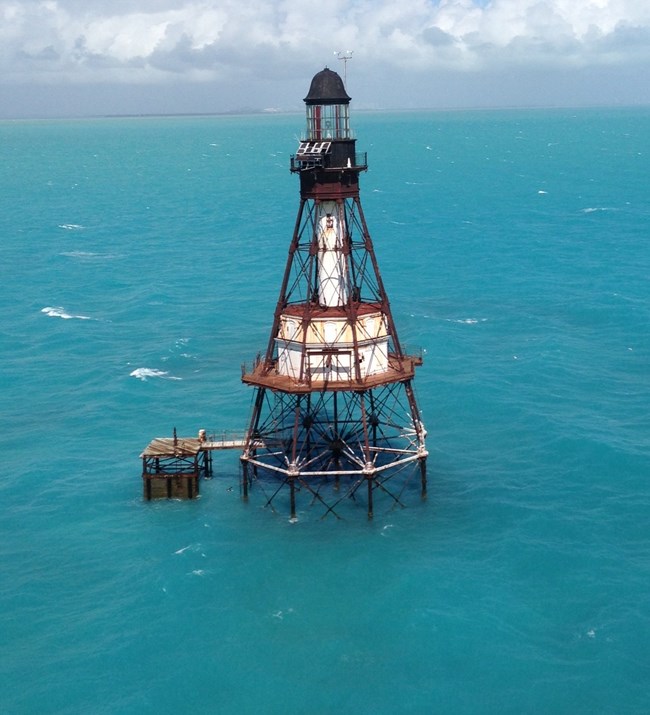 Aerial view of a rusty metal lighthouse rising above turquoise ocean waters