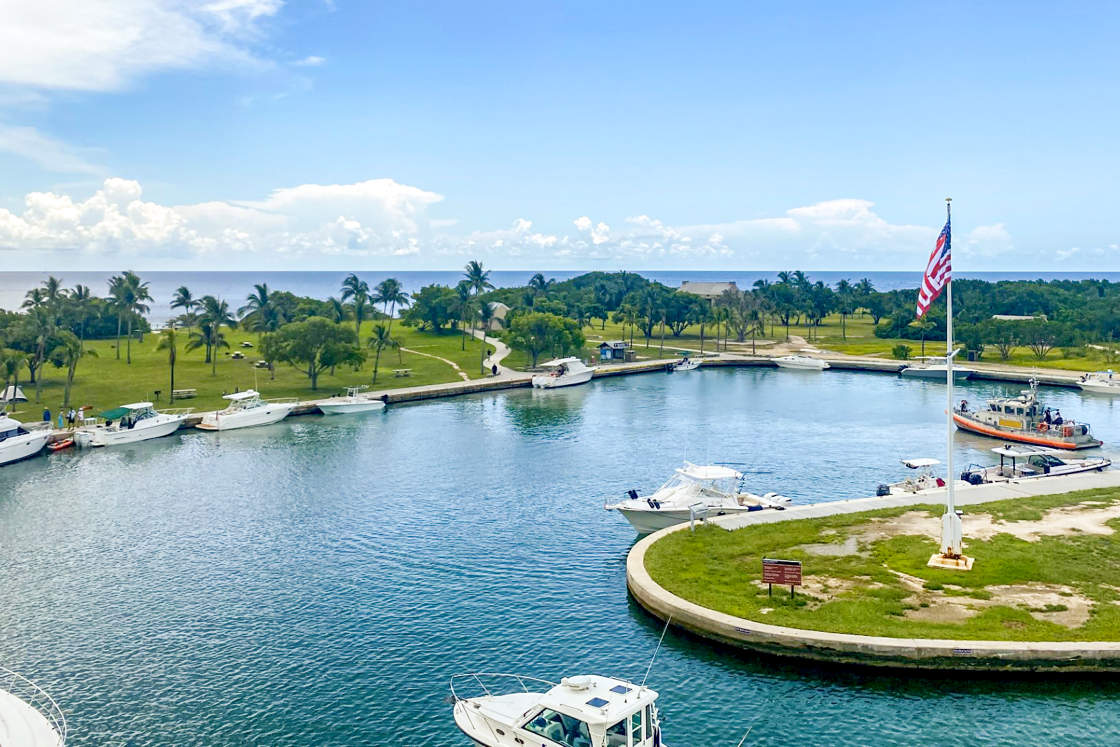 Boats are tied up to a sea wall. There is an American Flag, trees around the island, and blue ocean in the background.