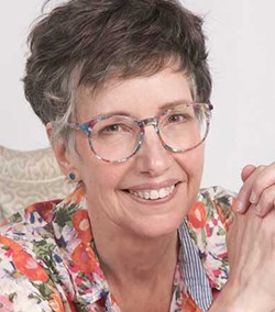 A smiling woman with short hair and colorful glasses wears a floral top and rests her hands together, posing against a light background.