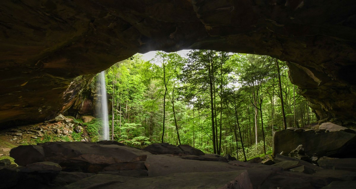 A white veil of water falls from a bluff above a bright green forest of trees are shown in beyond the rock arch that spans the front of the photo
