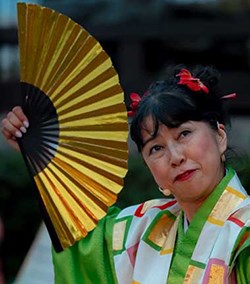 A woman in a colorful traditional Japanese kimono holds a gold and black fan, with red hair accessories and a headset mic, likely performing.