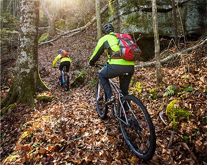 Two mountain bikers in bright yellow shirts riding up a leaf covered trail.