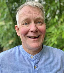 A smiling man in a light blue, buttoned shirt stands outdoors with green foliage in the background.