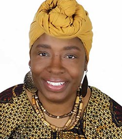 A smiling woman wearing a yellow headwrap, patterned clothing, large earrings, and beaded necklaces poses against a white background.