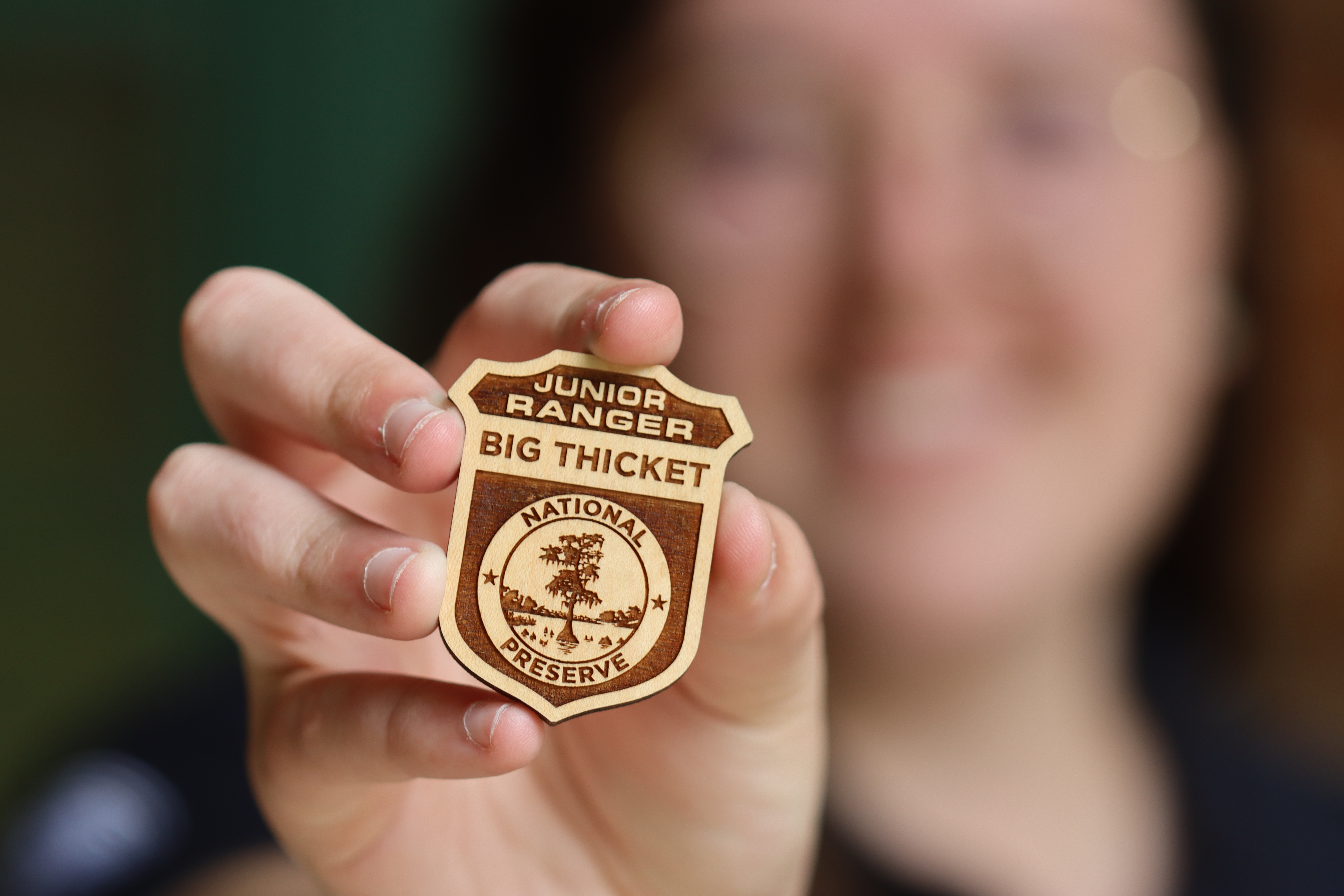 close-up of a hand holding a wooden junior ranger badge with text that reads Junior Ranger Big Thicket National Preserve, and an image of a bald cypress swamp.