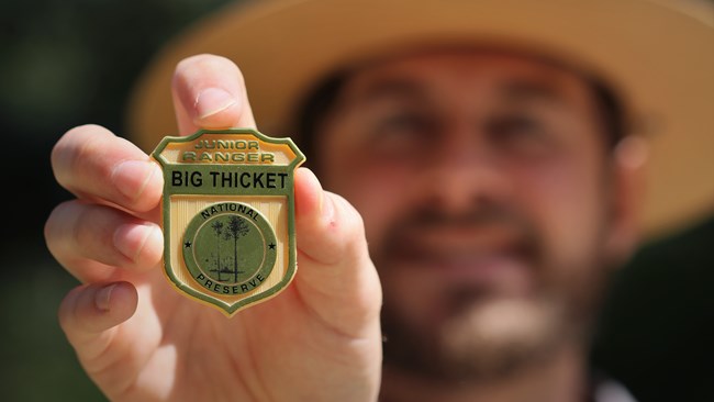 close-up of a park ranger holding a gold badge that reads "junior ranger; big thicket national preserve". A park ranger is out of focus in the background.