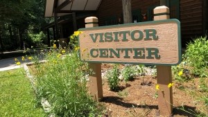 Wooden horizontal sign, reading Visitor Center, sits on two wooden posts about 2 feet off the ground amidst a garden of yellow flowers.