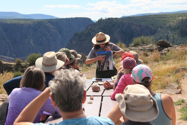 Park ranger in uniform holds a laminated paper. Visitors look at it from picnic table seating.