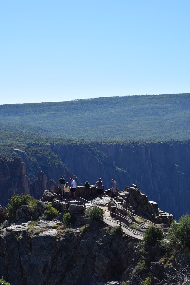 Rock outcropping with a path and metal railing leading to an overlook. Canyon and mesa are visible in background.