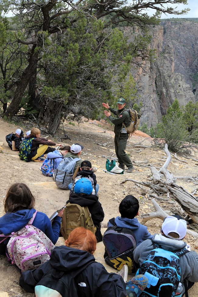 A park ranger stands in front of a group of schoolchildren. The children sit on a dirt path with backpacks and clipboards by them. Trees surround the path. A canyon is in the background.