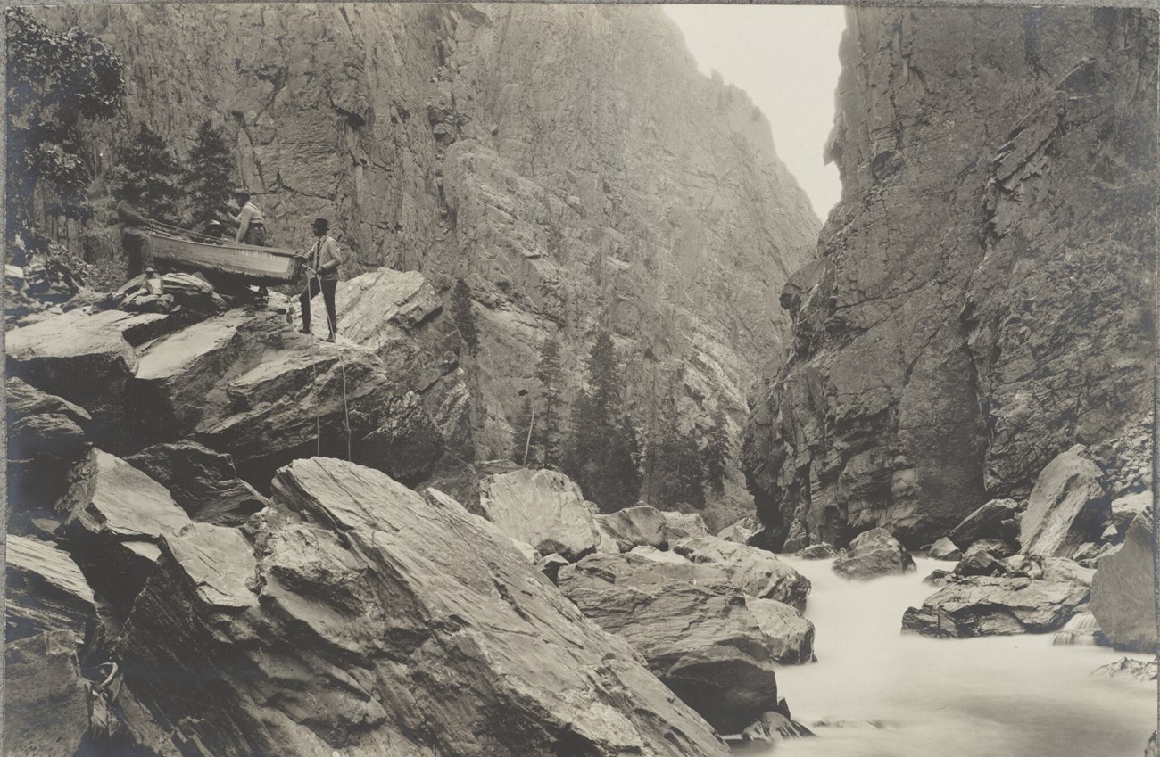 Men portage a wooden rowboat over boulders in the Black Canyon of the Gunnison. Gunnison River rapids are beside the rocks. Canyon walls rise around them.