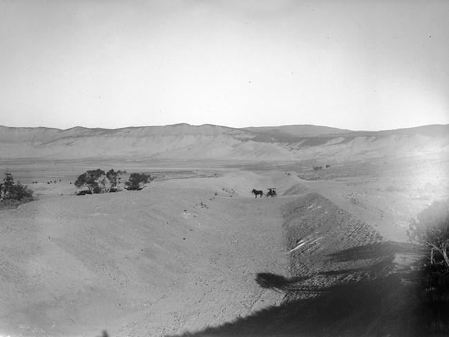 People in a horse-drawn carriage pass over a portion of a partially constructed and dry canal. Mountains are in the background.