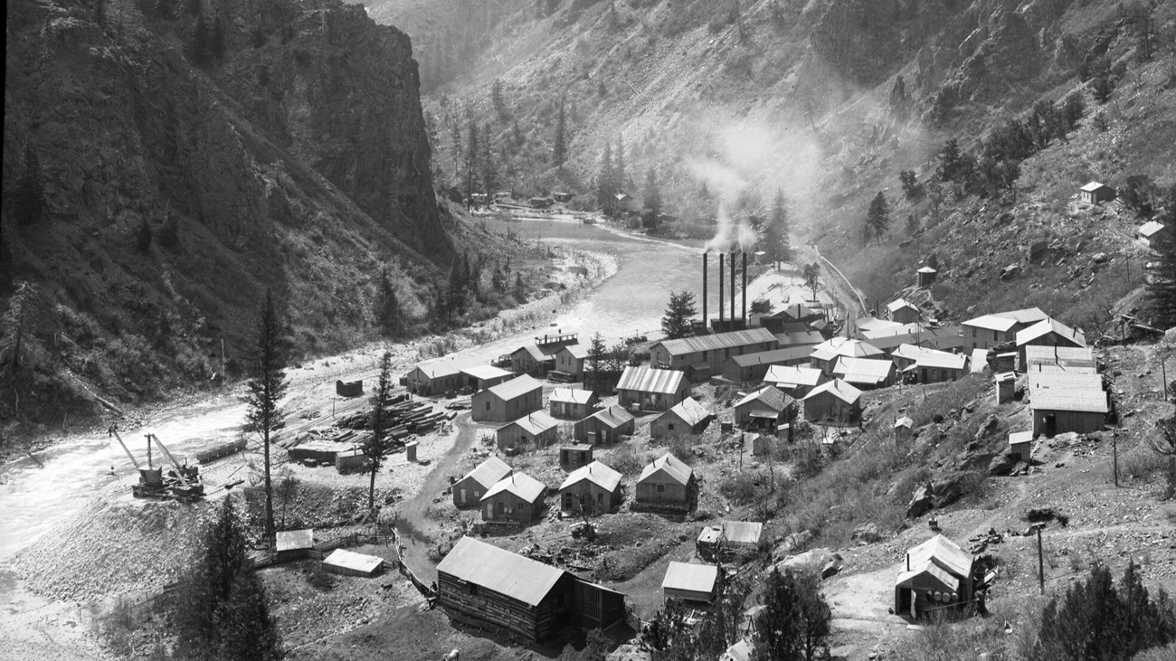 Historic image of the Gunnison River and East Portal. Workshops, a powerhouse, and other frame buildings are near ore cars, piles of timber, and railroad tracks. A river between canyon walls runs by the town.