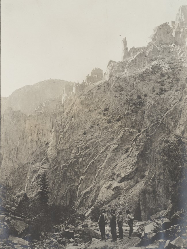 Historic image in sepia of a steep canyon. Four men stand pointing at cliffs and canyon walls around them.