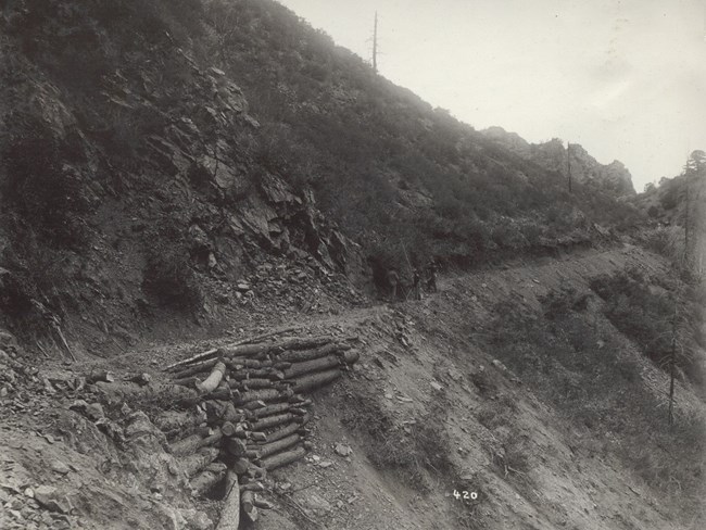 Historic image of three men working on road construction. The road is cut into the side of a canyon. Log reinforcements are visible under the road cut. Loose rock is above the roadway.