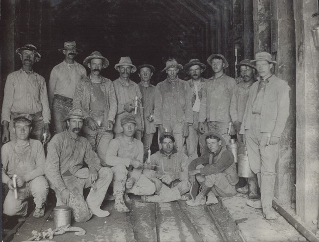 Historic image of a group of workers wearing hats and holding candles. A wooden lined tunnel is visible around them.