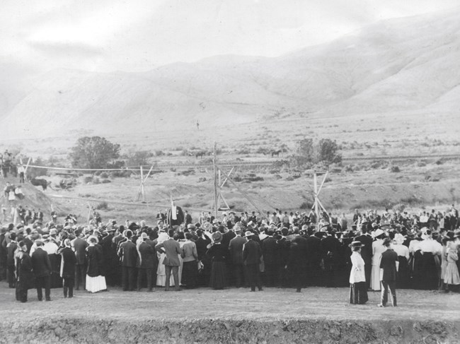 Historic photo of a crowd of people wearing clothes of the early 20th century. They stand near a canal ditch. Mesas are in the background.