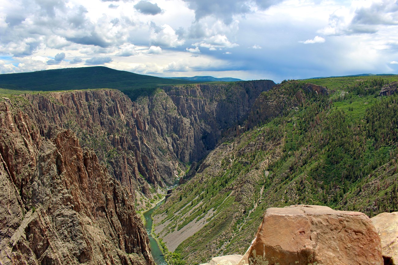 View of a deep canyon with dark walls. Green foliage is visible on the right side (north facing) canyon walls. A blue river is at the bottom. Blue sky and clouds above.