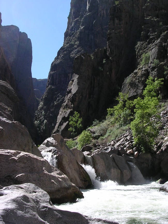 Steep dark canyon with large grey boulders and a river flowing. A small waterfall cascades over the rocks. Green vegetation is along the riverbank.