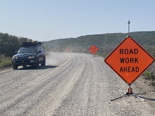 An orange diamond shaped sign with 'Road Work' sits on a gravel roadway. A dark colored vehicle drives by the sign in the other lane.