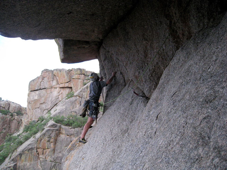 a rock climber clings to a gray rock wall underneath a large overhang