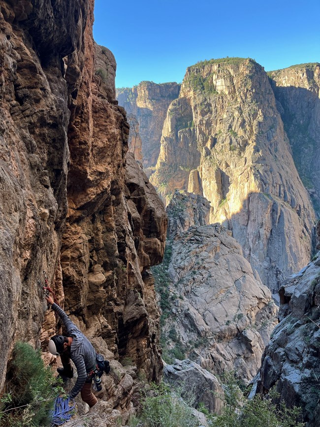 A person wearing a helmet and climbing gear stands near the edge of a vertical cliff. Canyon walls are in the background.