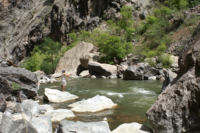 A person fishing while standing on a boulder next to a river