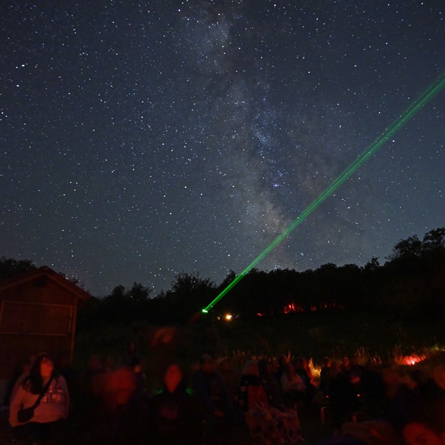 A crowd of people illuminated by red light look up at the sky. A green laser is pointing at the sky. The Milky Way is visible above the crowd.