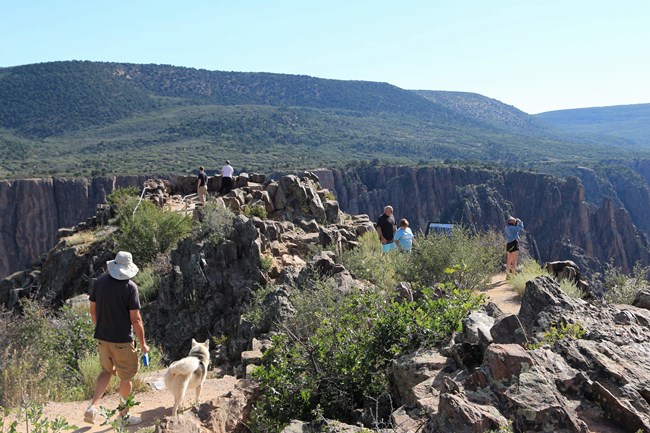 People hiking to a rocky overlook area. A person has a leashed dog in the foreground.