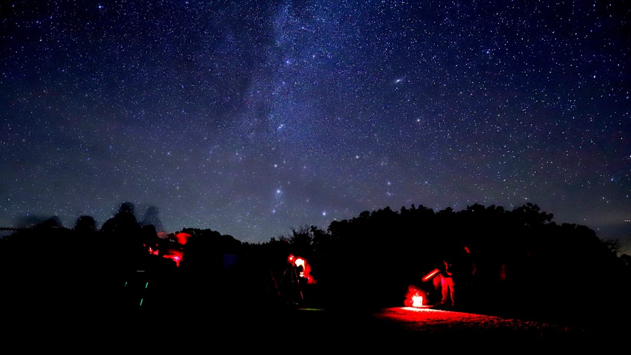 Milky Way over a dark scene with people using red light near telescopes