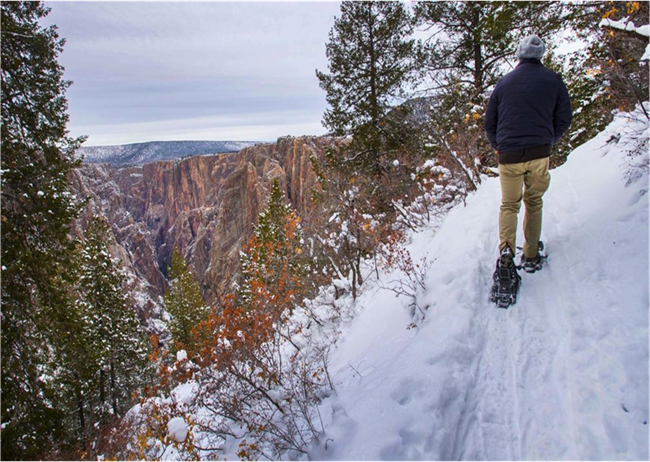 Snowshoer hikes along tree-lined trail with views of canyon.