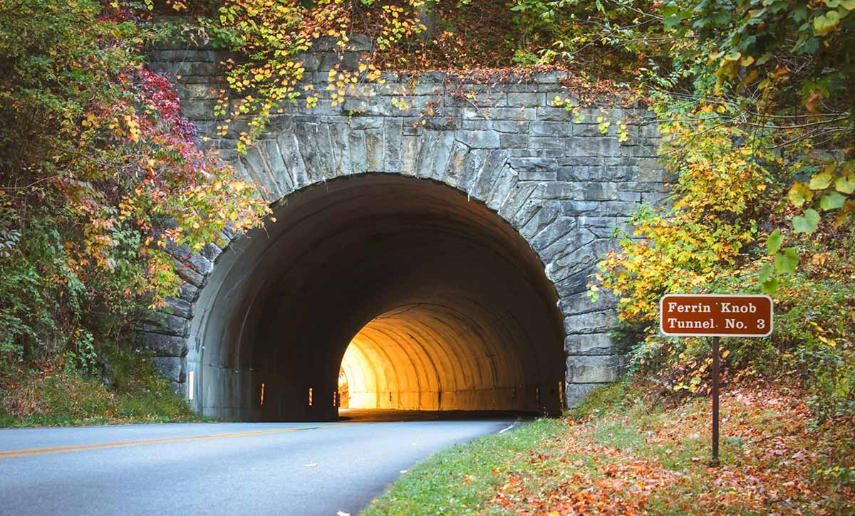 Sunlight illuminates the far end of a stone tunnel on the parkway. Red and yellow fall foliage surrounds the tunnel entrance.