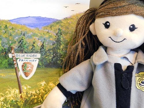 A rag doll dressed like a female park ranger stands next to a painting of a field with a Blue Ridge Parkway sign