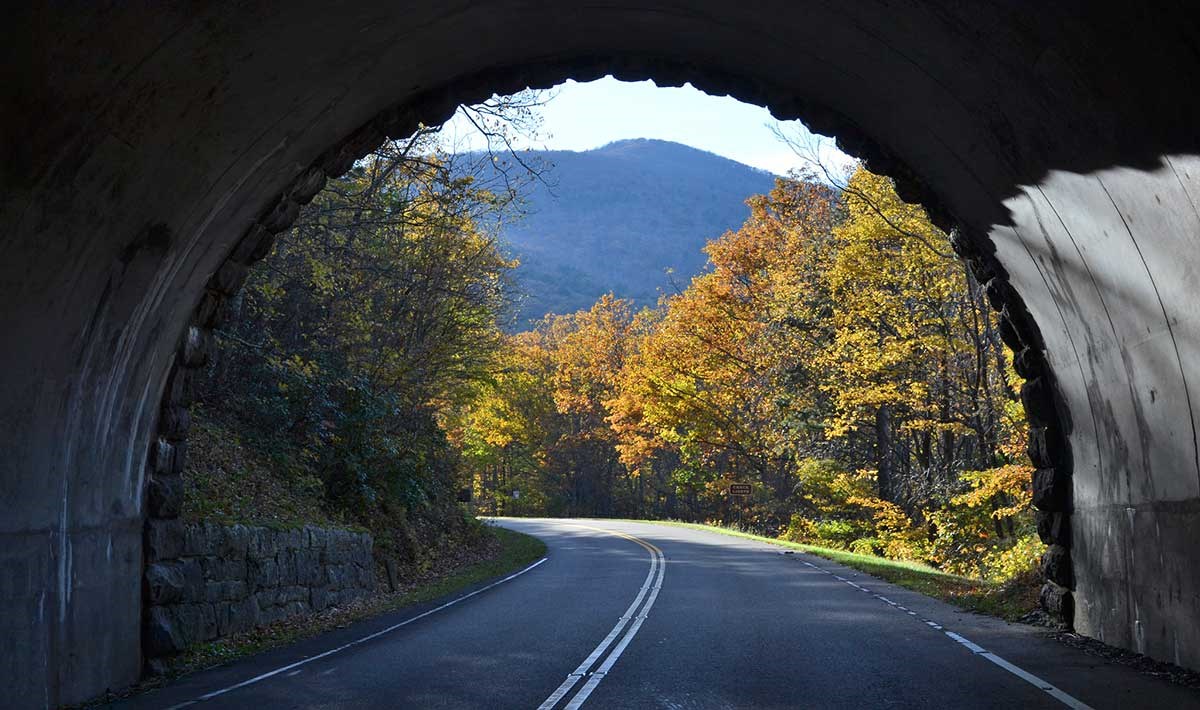Looking out of a tunnel on the parkway. The sides of the tunnel frame a view of a mountain in the distance and trees cloaked in fall colors lining the roadway.