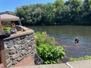 Stone wall alongside river. IN the river, a scuba diver stands holding a hose. On the shore, a white tent with people standing under it