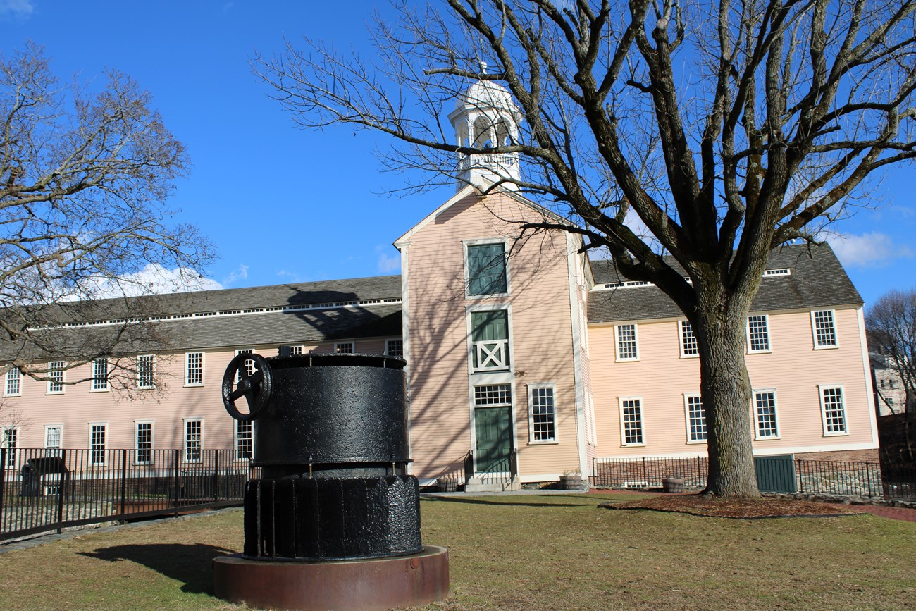 Yellow two-story mill with three story bell tower. A large black object called a turbine is on the lawn in front of the building