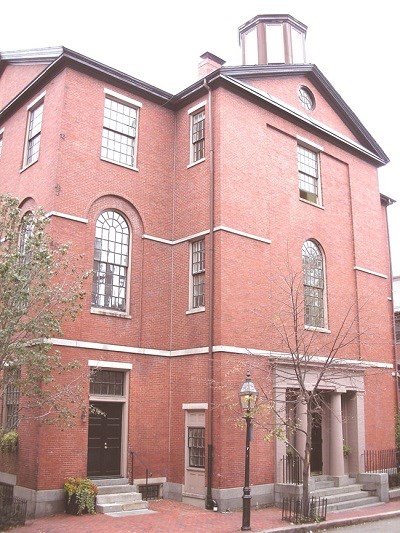 four story brick building with arched windows on the second story. Façade slightly juts out in the center entryway.