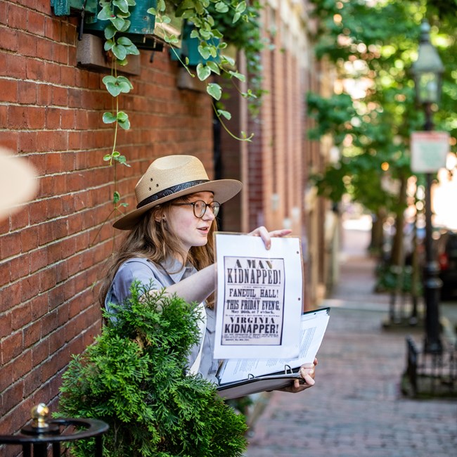 A ranger holds a flier while standing near a brick wall under a flowerbox