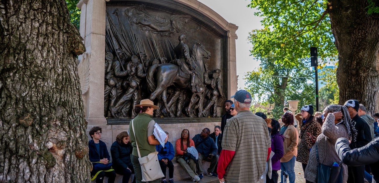 A ranger faces a crowd while standing in front of a large monument