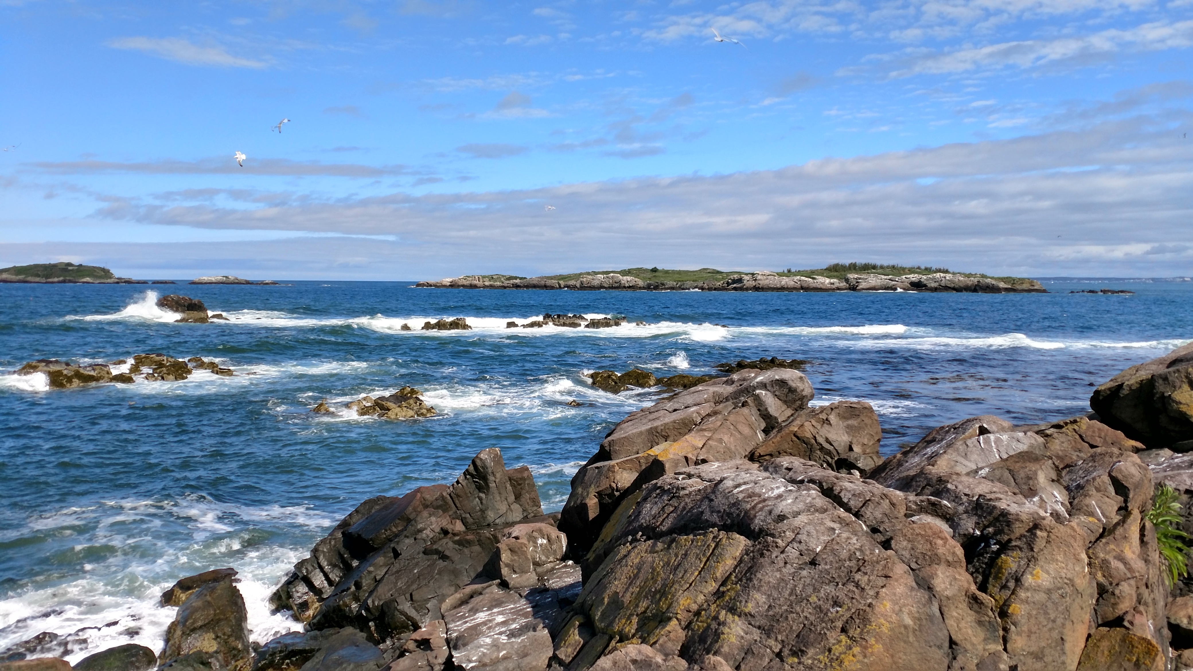 reddish brown rocky shore juts out into the deep blue waters. Three islands in the distance