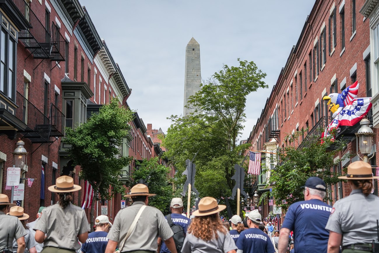 A group of uniformed rangers and volunteers walk in a parade with a tall monument in front of them
