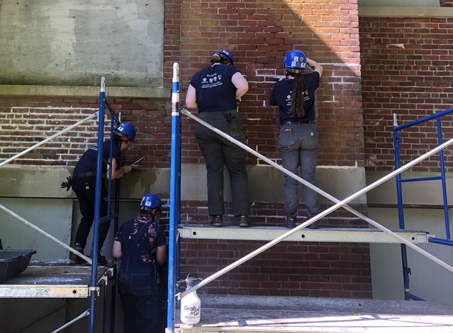 Four crew members wearing hard hats in front of an old brick wall. Three of them are on scaffolding, and one is standing on the ground below. The mortar in several parts of the wall is much brighter white than the mortar on the rest of the wall.