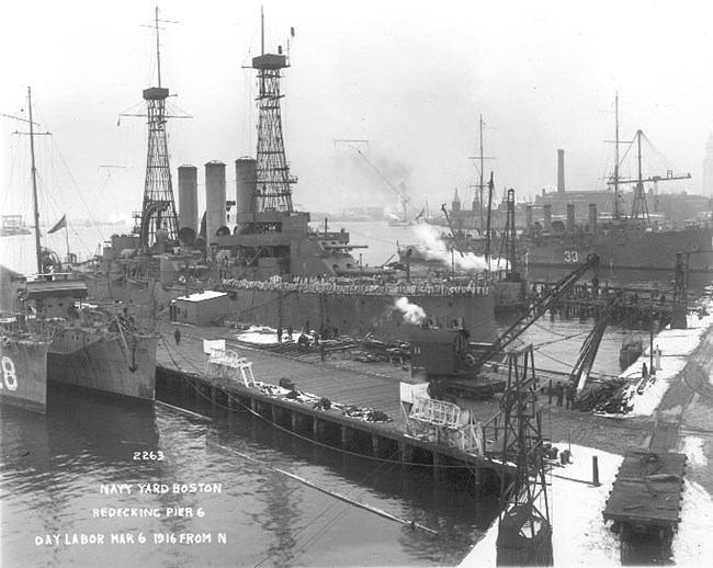 Photograph of warships of different classes moored to the wooden pier 6. Snow is covering parts of the ground.