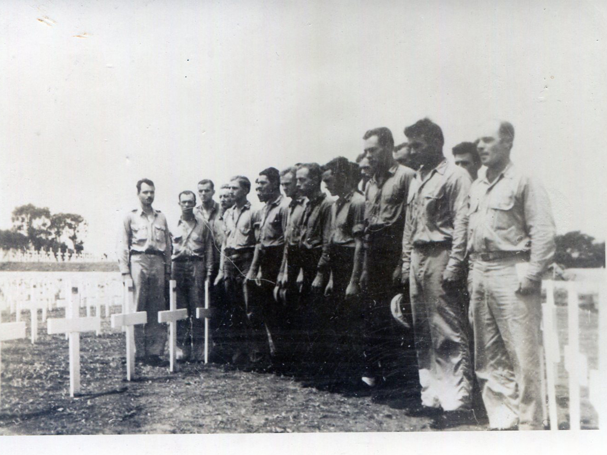 Group of Navy Sailors standing side-by-side in a cemetery. They all have their heads bowed down.